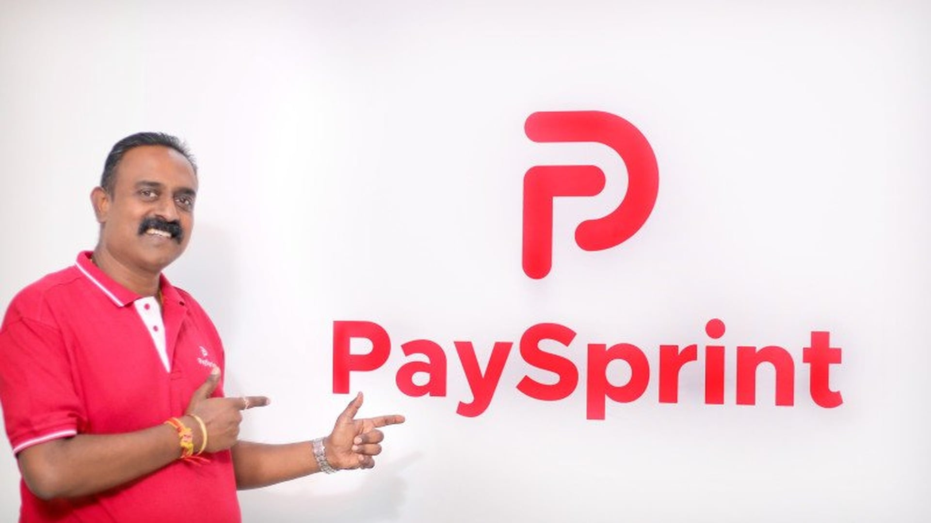 PaySprint founder and CEO S. Anand in a red polo shirt pointing at the PaySprint logo on a white wall
